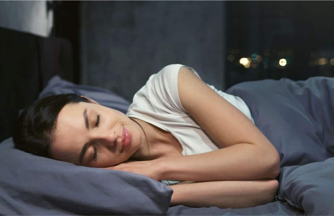Eine Frau mit dunklen Haaren liegt schlafend in einem Bett. Die Bettwäsche ist dunkelgrau.