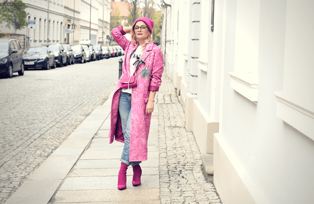 Eine Frau trägt einen Mantel, Schuhe, eine Handtasche und eine Mütze in Pink. Dazu hat sie eine blaue Jeans und ein weißes T-Shirt an. Sie steht auf einer Straße im Hintergrund sieht man Hauseingänge und parkende Autos.