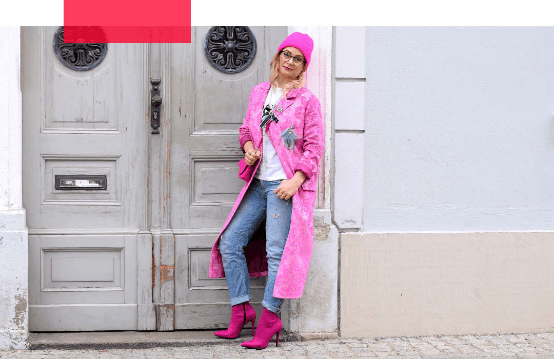 Eine Frau mit blonden, langen Haaren und Brille lehnt in lässiger Pose mit dem Rücken an einem Hauseingang. Sie trägt einen Brokatmantel, eine Mütze, eine Tasche und Schuhe in Pink. Dazu hat sie eine Jeans und ein helles T-Shirt kombiniert.