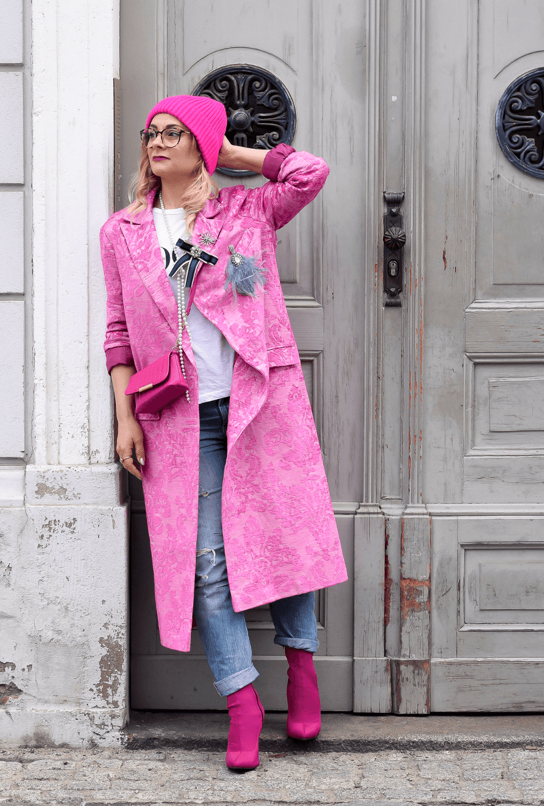 Eine Frau trägt einen Mantel, Schuhe, eine Handtasche und eine Mütze in Pink. Dazu hat sie eine blaue Jeans und ein weißes T-Shirt an. Sie steht vor einem Hauseingang mit einer grauen Holztür.
