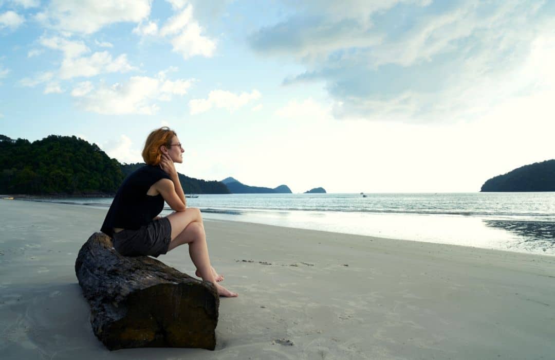 Eine Frau sitzt auf einem Baumstand der am Strand liegt und schaut auf das Meer. 