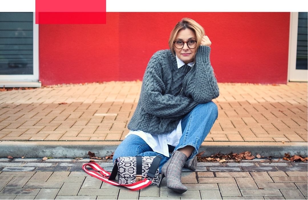 Blonde Frau mit Brille sitzt vor einer roten Wand. Sie trägt einen grauen Pullover über einem weißen Hemd und eine hellblaue Jeans.