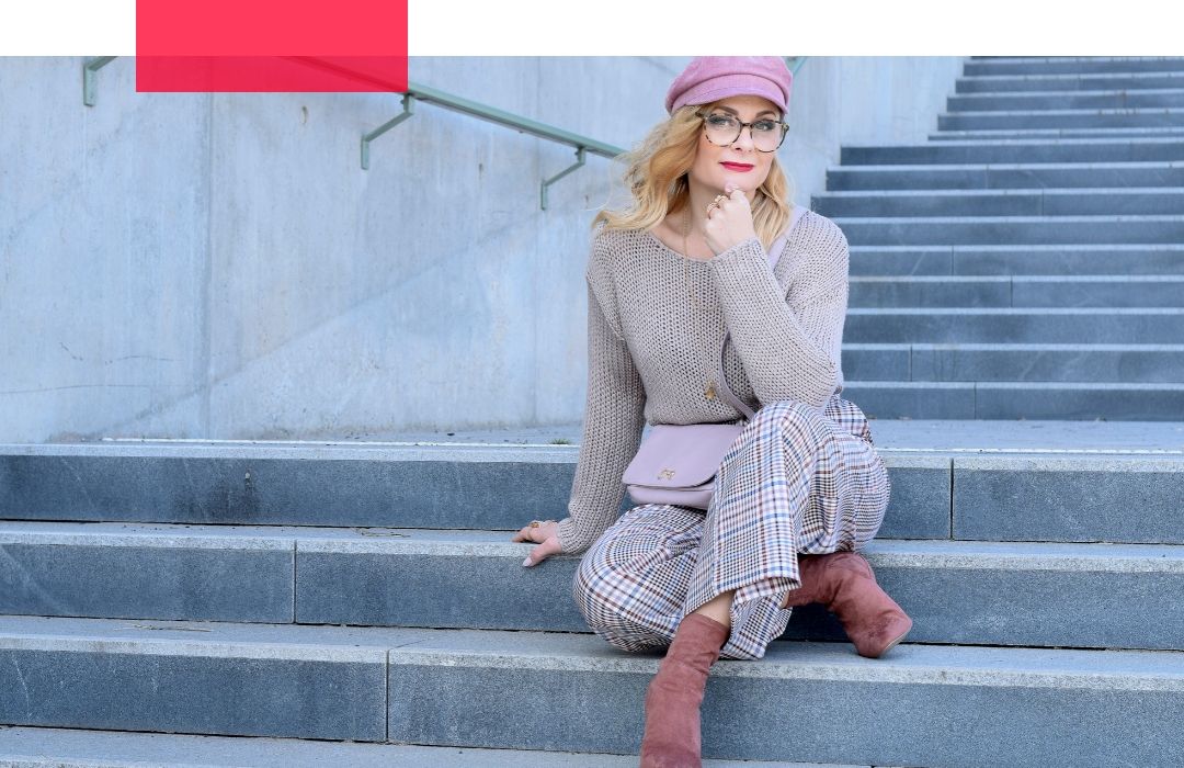 Frau mit langen, blonden, gelockten Haaren sitzt auf einer Treppe. Sie trägt eine Beige, Grau, Rosa karierte Culotte und eine rosane Bakerboy Mütze.