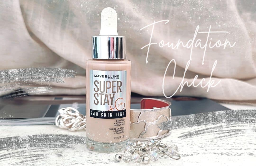 Eine Flasche des Maybelline 24h Super Stay Skin Tint Foundation steht auf einem Tisch. Darum liegt Silberschmuck.