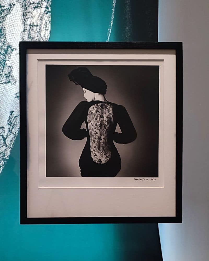 Exponat der Yves Saint Laurent Ausstellung in Calais. Bild von einer gerahmten Schwarz-Weiß Fotografie. Das Bild zeigt eine Frau in einem schwarzen Spitzenkleid von Yves Saint Laurent. Die Frau hat dem Betrachter den Rücken zugedreht. Der Rücken ist bedeckt mit einem schwarzen Spitzenstoff.