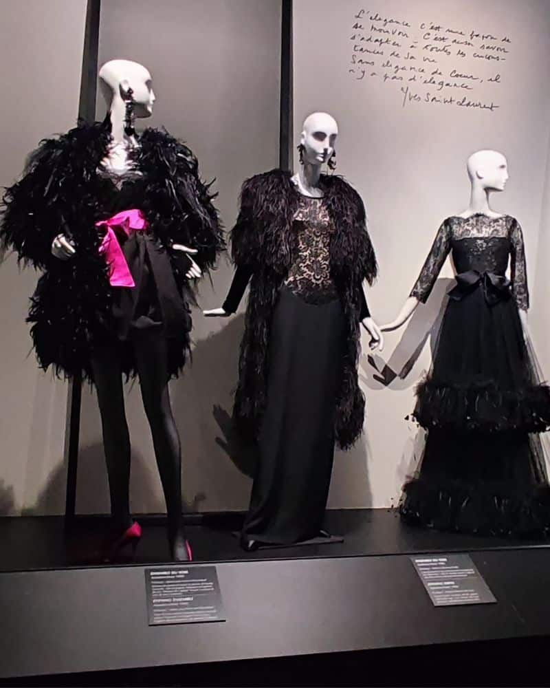 Drei Kreationen in der Yves Saint Laurent Ausstallung in Calais. Die Kleider sind auf weiße Schaufensterpuppen drapiert. Die Kleider sind alle schwarz, zwei sind lang und eines kurz. Das kurze Kleid hat eine Schleife in Pink in der Taille. Schwarze Spitze und schwarze Federn sind elementare Elemente in den Entwürfen.