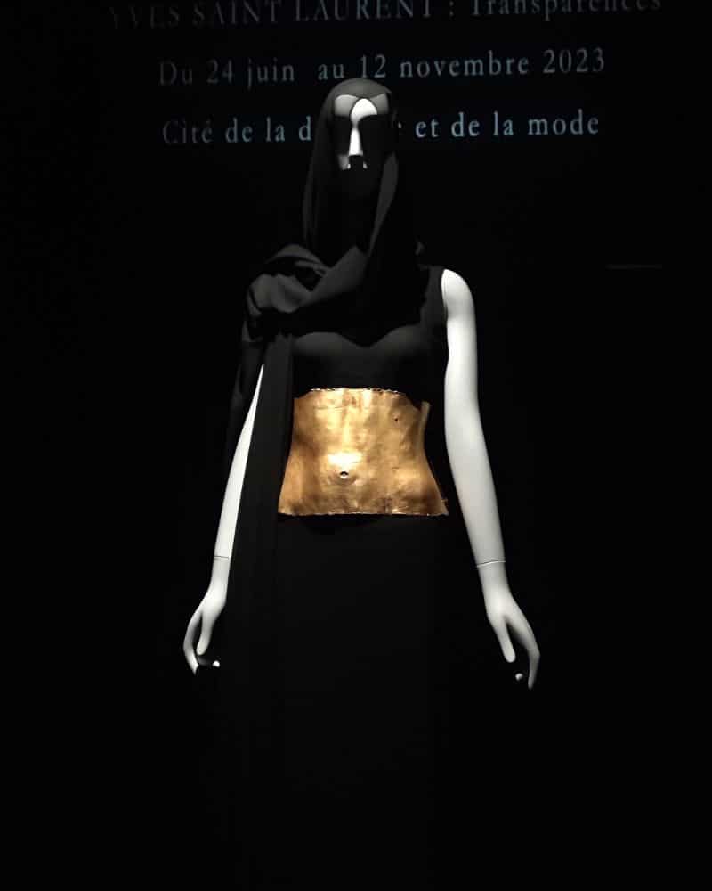 Ein Exponat der Yves Saint Laurent Ausstellung in Calais. Eine weiße Schaufensterpuppe komplett in einem schwarzen, kurzärmeligen Kleid mit breitem, goldenem Gürtel. Über den Kopf ist ein schwarzer Schleier drapiert.