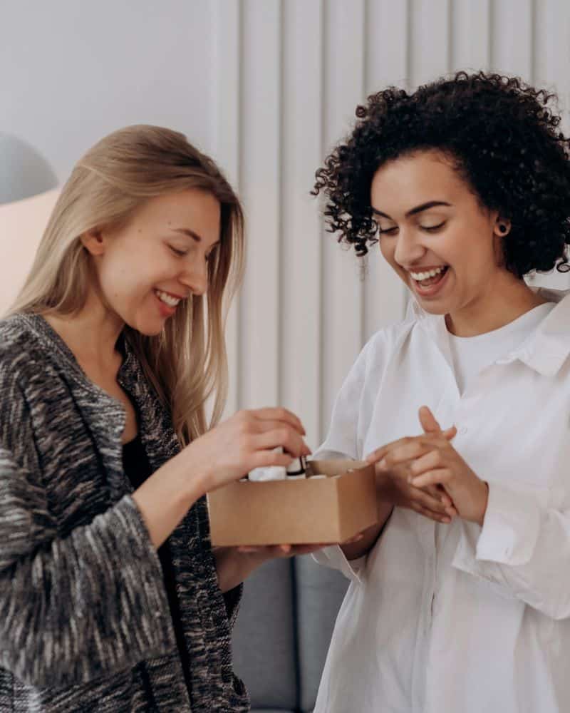 Anti-Aging Produkte. Zwei junge Frauen stehen in einem Raum und haben gemeinsam ein Päckchen mit unterschiedlichen Produkten in der Hand auf das sie blicken.