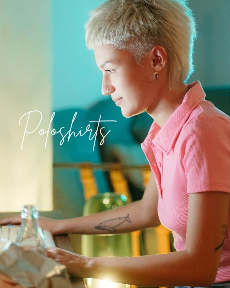 Frau mit kurzen blauen Haaren trägt ein rosa Polohemd und sitzt an einem Tisch. Modisches Polohemd für 2024.