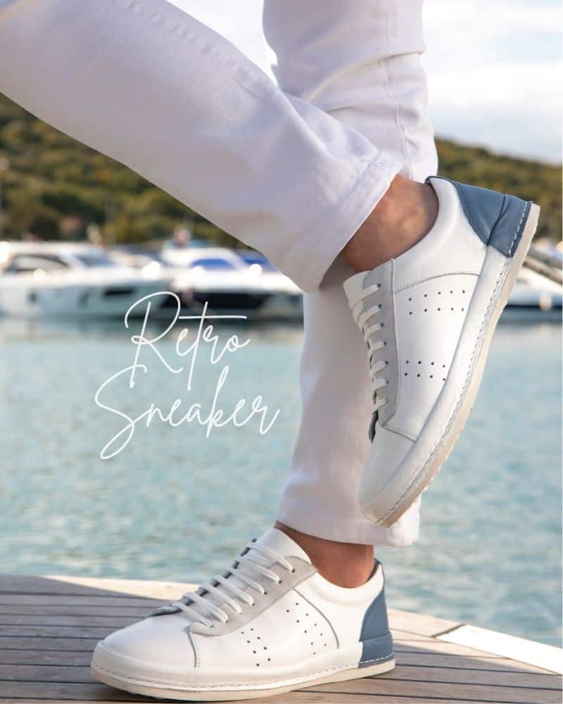 Nahaufnahme zweier Beine in weißen Hosen und hellen Sneakern mit schmalem Profil vor einem Hafen mit blauem Meer. Retro Sneaker Modetrend 2024.