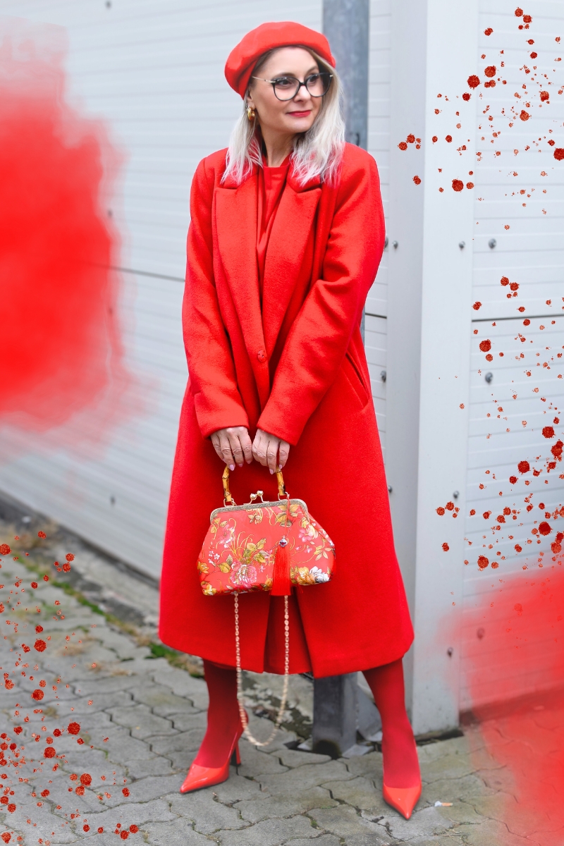 Blonde Frau steht an einer Wand und trägt ein Allover Rot Outfit. Der Look besteht aus: einem roten Kleid, einem roten Mantel plus roter Strumpfhose zu roten Pumps. Die Accessoires sind eine rote Tasche und eine rote Baskenmütze.