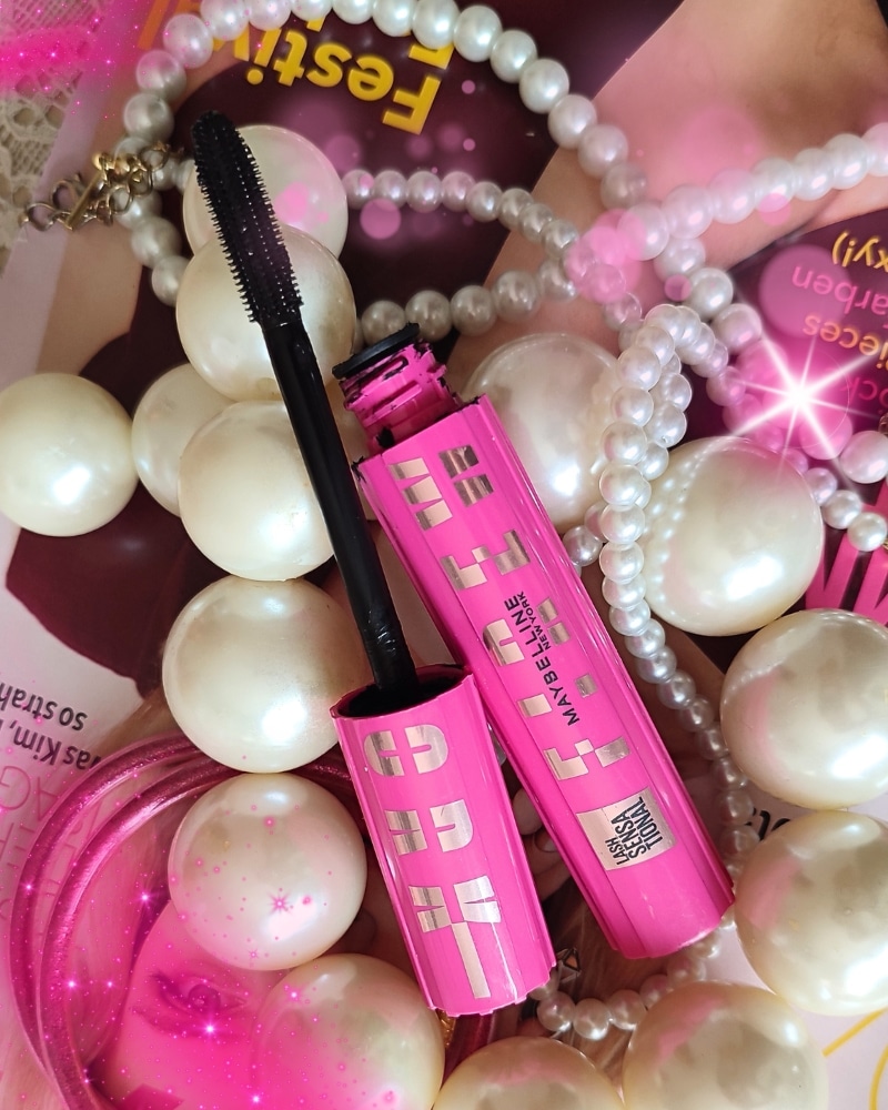 Die pinke Maybelline Firework Mascara liegt geöffnet zusammen mit einigen Perlenketten auf einem Tisch.